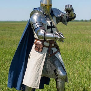 May include: A full suit of medieval knight armor, including a helmet with a gold visor, shoulder plates, and chainmail. The knight is holding a sword and wearing a white tunic with a black cross and a blue cape. The scene is set in a grassy field.
