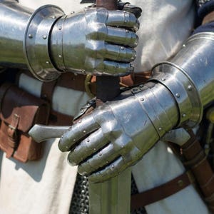 May include: Close-up of a medieval knight's gauntlets holding a sword. The gauntlets are made of polished steel and are riveted. The sword has a brown leather-wrapped handle. The knight is wearing a white tunic with a black cross.