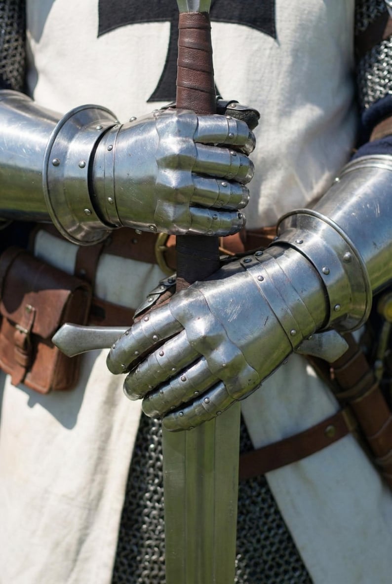 May include: Close-up of a medieval knight's gauntlets holding a sword. The gauntlets are made of polished steel and are riveted. The sword has a brown leather-wrapped handle. The knight is wearing a white tunic with a black cross.