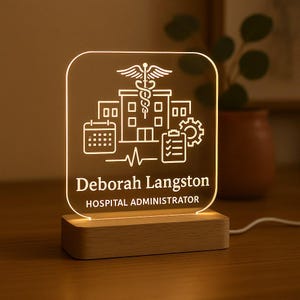 Może przedstawiać: Spersonalizowana lampa biurkowa z przezroczystym panelem akrylowym z symbolami medycznymi i ikonami szpitala, imieniem "Deborah Langston" i tytułem "HOSPITAL ADMINISTRATOR". Lampa stoi na drewnianej podstawie i jest podświetlana.