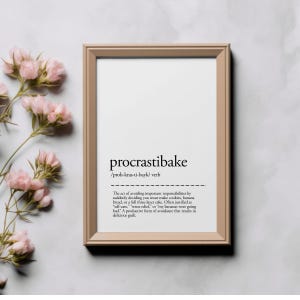 May include: A framed print with the word "procrastibake" in a modern font. The definition below humorously describes avoiding responsibilities by baking. Pink flowers are arranged near the frame.