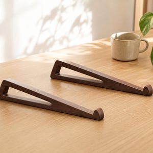 Peut inclure: Une paire de supports pour ordinateur portable en bois brun foncé, présentant une conception triangulaire à cadre ouvert avec un petit crochet incurvé vers le haut à l'avant pour maintenir l'appareil. Ils ont une finition lisse et mate.