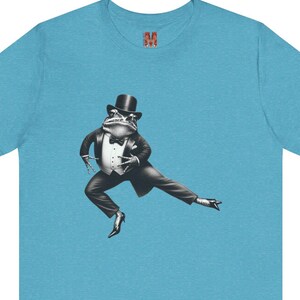 Könnte beinhalten: Ein hellblaues T-Shirt mit kurzen Ärmeln und einer Grafik eines Frosches, der einen schwarzen Smoking, einen Zylinder und eine Fliege trägt und eine Tanzpose einnimmt. Der Frosch ist in Schwarz-Weiß gehalten. Ein kleines rotes M ist im Nackenbereich gedruckt.