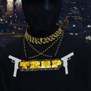 May include: Black t-shirt with the word "TRAP" in yellow letters, framed by two white pistols. The person is wearing a black mask and multiple gold chain necklaces. The background shows a city at night.