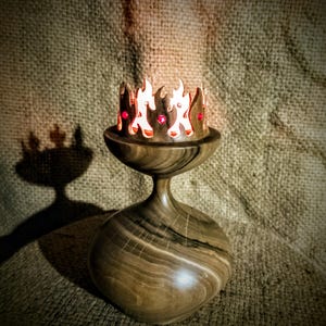 Puede incluir: Un candelabro de madera hecho a mano con una corona en forma de llama. El candelabro es de madera oscura con un acabado liso. La corona tiene detalles rojos y está iluminada, proyectando una sombra. El diseño recuerda a una corona.