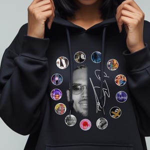 May include: Black hoodie featuring a grayscale image of a person wearing glasses, surrounded by circular images. The hoodie also displays a signature in white. The design is centered on the front of the garment.