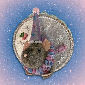May include: A whimsical, plush mouse wearing a party hat and ruffled collar, set against a glittery, circular backdrop. The mouse has tan ears, a pink nose, and is adorned with decorative elements like stars and a heart.