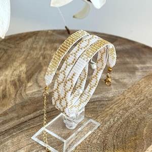 Könnte beinhalten: Ein gewebtes Perlenarmband mit einem sich wiederholenden geometrischen Kettengliedmuster in Weiß und Metallic-Gold. Das Armband verfügt über einen goldfarbenen Karabinerverschluss, eine kurze Verlängerungskette und kleine weiße Kugelperlen.