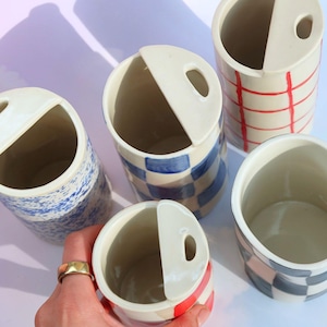 Könnte beinhalten: Eine Sammlung von Keramikbehältern mit einem einzigartigen Design. Die Behälter weisen verschiedene Muster auf, darunter blaue Spritzer, Schachbrettmuster und rote Gitterlinien. Jeder Behälter hat einen weißen Deckel mit einem kleinen Loch. Die Behälter sind zylindrisch und scheinen handgefertigt zu sein.