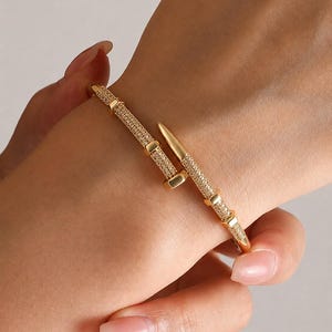 May include: A gold-toned bangle bracelet shaped like a bent nail, featuring a textured surface encrusted with numerous small, clear, sparkling stones. The design includes a pointed end and a flat head, creating a continuous, wrap-around appearance.