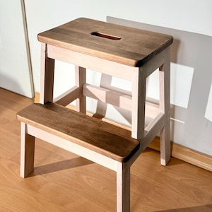 Puede incluir: Un taburete de madera de dos escalones con una parte superior rectangular y un escalón inferior. La parte superior y el escalón están teñidos de marrón oscuro, mientras que las patas y el marco son de color madera clara. Un asa está cortada en la parte superior.