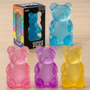 Op de afbeelding: Een verzameling doorschijnende gummy bear-speelgoed in verschillende kleuren, waaronder blauw, roze, paars en geel. Eén blauwe beer wordt getoond naast een productdoos met de woorden "Gummy Bear" en "Simpitte".