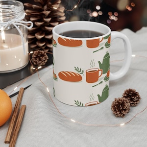 May include: White ceramic mug with a repeating pattern of bread loaves, teacups, teapots, and sprigs in orange and green. The mug is filled with a dark liquid and has a white handle.