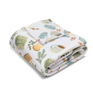 May include: A folded white blanket featuring a repeating pattern of winter-themed illustrations. The design includes mugs, mittens, scarves, and foliage in green, brown, and blue hues.