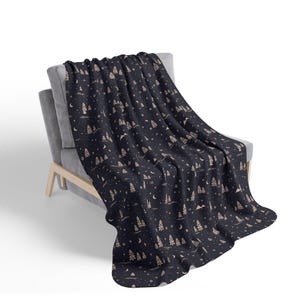 May include: A dark blue throw blanket draped over a gray armchair. The blanket features a repeating pattern of beige trees, mountains, moons, and stars. The blanket is made of a soft material and is suitable for a living room or bedroom.