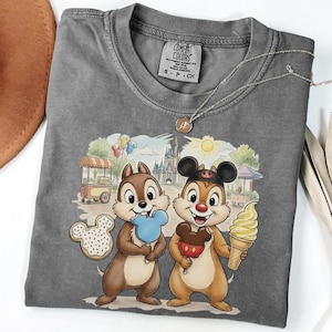 May include: Gray Comfort Colors t-shirt featuring Chip and Dale holding treats in front of a Disney park scene. The shirt has a crew neck and short sleeves. A gold necklace is draped across the shirt.