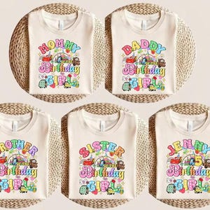 May include: Five beige t-shirts with colorful graphics for a birthday celebration. The shirts feature text like "Mommy," "Daddy," "Brother," "Sister," and a name, with the words "of the Birthday Girl" and various cartoon vehicles.