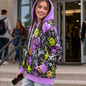 Peut inclure: Un sweat-shirt à capuche à manches longues avec un imprimé intégral vibrant sur fond noir. Le motif comprend des visages souriants jaunes, des cœurs violets et argentés, des étoiles et des notes de musique. Capuche, poignets et ourlet violet clair.