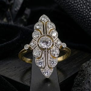 May include: A vintage-style ring with a gold band and a central cluster of diamonds. The ring features a detailed, ornate design with multiple smaller diamonds surrounding a larger, central stone. The setting is silver-toned, and the ring is displayed against a dark background.
