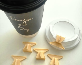 May include: A black paper coffee cup with a lid, and the words "Energize your Day" printed on the side. Several beige plastic cup plugs are scattered around the cup and lid. The lid is black with a beige plastic plug attached.