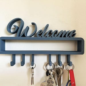 Puede incluir: Un llavero azul con la palabra "Welcome" en escritura cursiva. El llavero tiene un estante rectangular con seis ganchos debajo, cada uno con llaves. El llavero está montado en una pared blanca.