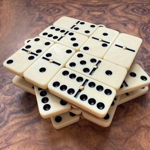 May include: Ivory-colored dominoes with black dots and gold-colored center pins are stacked on a wooden surface. The dominoes are arranged in a slightly haphazard pile, showcasing the game pieces.