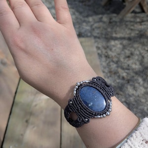 May include: A handmade macrame bracelet featuring a large, oval, blue gemstone. The dark gray macrame setting is accented with small silver beads. The bracelet is worn on a wrist.