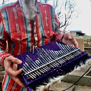 Peut inclure: Un kalimba, instrument de musique, est tenu devant une personne. Le kalimba a des dents métalliques et est posé sur une base violette à pois blancs. L'instrument est garni de fourrure blanche.