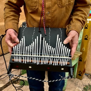May include: A black kalimba instrument with white tines and a wooden base. The instrument is held by a person. The tines are arranged in a curved pattern. The kalimba has a hole in the center and is connected to a black and white cord.