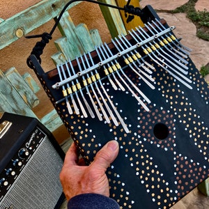 Può includere: Un kalimba nero con lamelle argentate e un motivo decorativo di punti bianchi, dorati e marroni. Lo strumento ha una cornice in legno e una tracolla nera. Un amplificatore è visibile sullo sfondo.