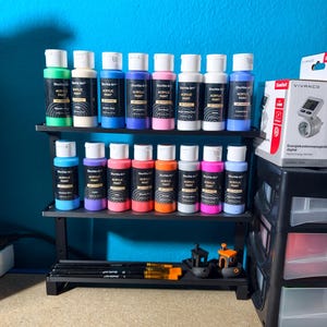 May include: A black metal shelf displays various bottles of Shuttle Art acrylic paint in a range of colors, including blue, pink, and white. Paintbrushes and small 3D-printed models are also visible, suggesting a crafting or art supplies theme.
