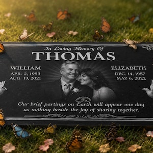 Op de afbeelding: Zwarte granieten gedenkplaat met de naam "THOMAS" bovenaan, met foto's van een stel. De plaat bevat data en een citaat, omringd door vlinders, duiven en rozen.