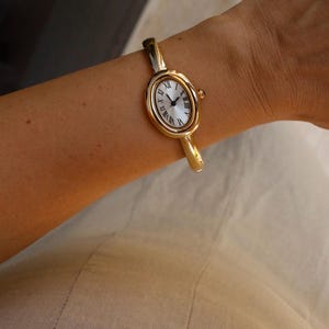 Può includere: Orologio da polso ovale dorato con quadrante bianco e numeri romani neri. L'orologio ha un cinturino dorato. L'orologio è indossato al polso.