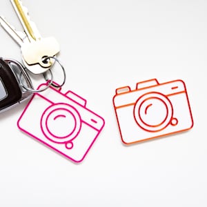 May include: A set of two camera-shaped keychains, one in bright pink and one in orange, alongside a silver metal car key fob and a standard house key attached to a circular metal ring, all displayed against a plain white background.