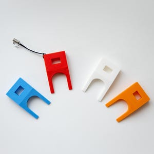 May include: Four colorful, 3D-printed keychains in red, blue, white, and orange. Each keychain has a square cutout and a unique shape with two curved prongs. One red keychain has a black lanyard attached.