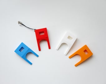 May include: Four colorful, 3D-printed keychains in red, blue, white, and orange. Each keychain has a square cutout and a unique shape with two curved prongs. One red keychain has a black lanyard attached.
