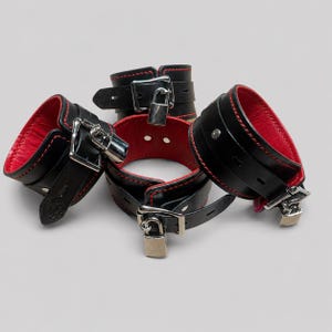 Puede incluir: Restricciones de muñeca de cuero negro con forro y costuras rojas. Cada restricción presenta una hebilla plateada y un candado para mayor seguridad. Diseñado para uso adulto.