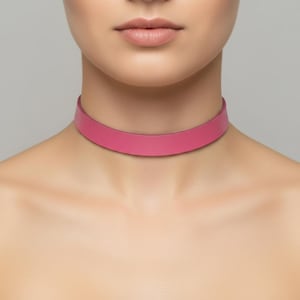 May include: A vibrant pink choker necklace. The necklace is a simple band of solid color, resting around the neck. The background is a neutral gray, highlighting the necklace's bold color and minimalist design.