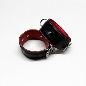 May include: Black leather wrist restraints with red interior lining. The restraints have silver-colored metal buckles and a D-ring. The edges are stitched with red thread. The wrist restraints are on a white background.