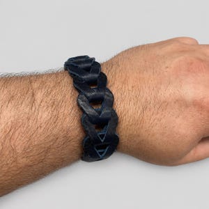 May include: A dark navy blue leather bracelet featuring a series of interconnected, braided-style links. The bracelet has a matte texture and a wide, structured design that wraps around the wrist, showcasing a repeating geometric pattern.
