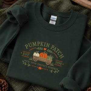 May include: A dark forest green crewneck sweatshirt featuring an embroidered graphic of a wooden wagon filled with three pumpkins. The text reads PUMPKIN PATCH, EST 1998, and PICK YOUR PERFECT PUMPKIN, accented with small orange hearts and leaves.