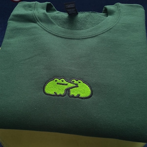 May include: Dark green sweatshirt with a crew neck. The sweatshirt features an embroidered design of two green frogs with black outlines. The frogs are positioned close together. The sweatshirt is folded neatly.