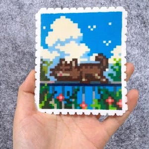 Puede incluir: Un dise&ntilde;o de arte pixelado que presenta un gato marr&oacute;n descansando sobre una repisa, sobre un cielo azul con nubes blancas. La obra de arte est&aacute; enmarcada con un borde blanco festoneado, que se asemeja a un sello. El dise&ntilde;o incluye follaje verde y flores rojas.