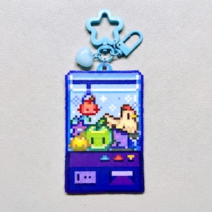 May include: A blue pixelated keychain with a claw machine design, containing pixelated objects. The keychain has a blue star-shaped clasp and a heart charm. The claw machine has a purple base and a blue top.