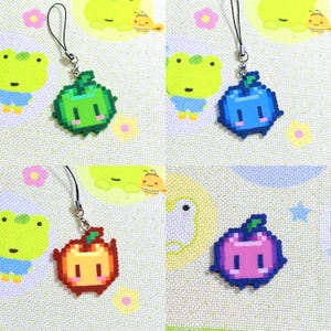 May include: Four pixel art fruit charms: a green apple, a blue blueberry, an orange apple, and a purple plum. Each charm has a black lanyard attached. The charms are on a patterned background with cartoon frogs and other designs.