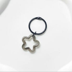 Puede incluir: Un anillo de metal negro con un dije de flor de cinco p&eacute;talos gris claro y transparente. El dije est&aacute; suspendido del anillo por un peque&ntilde;o conector negro. El anillo y el dije est&aacute;n sobre un fondo blanco.