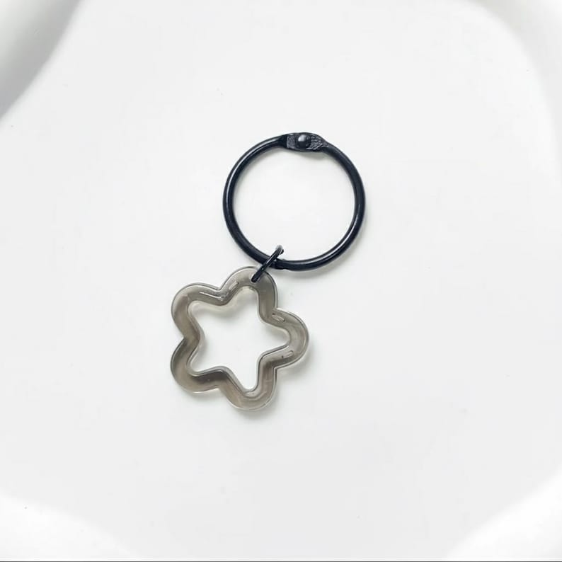 Puede incluir: Un anillo de metal negro con un dije de flor de cinco p&eacute;talos gris claro y transparente. El dije est&aacute; suspendido del anillo por un peque&ntilde;o conector negro. El anillo y el dije est&aacute;n sobre un fondo blanco.