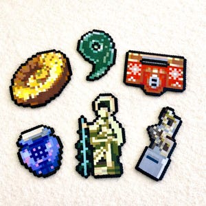 May include: A collection of pixel art-style magnets. The magnets include a donut, the number nine, a boombox, a blue jar, a figure with a staff, and a silver object. The magnets are made with a variety of colors.