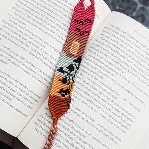 Puede incluir: Marcapáginas hecho a mano con un diseño tejido colorido. El marcapáginas presenta una parte superior roja con un motivo de casa y pájaro, que pasa a patrones geométricos naranjas, marrones y negros. Una borla trenzada añade un toque decorativo. Está colocado en un libro abierto.