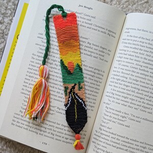 Puede incluir: Marcapáginas hecho a mano con un diseño tejido colorido. El marcapáginas presenta un degradado de naranja, amarillo, verde y negro, con forma de hoja en la parte inferior y una borla. Está colocado en un libro abierto.